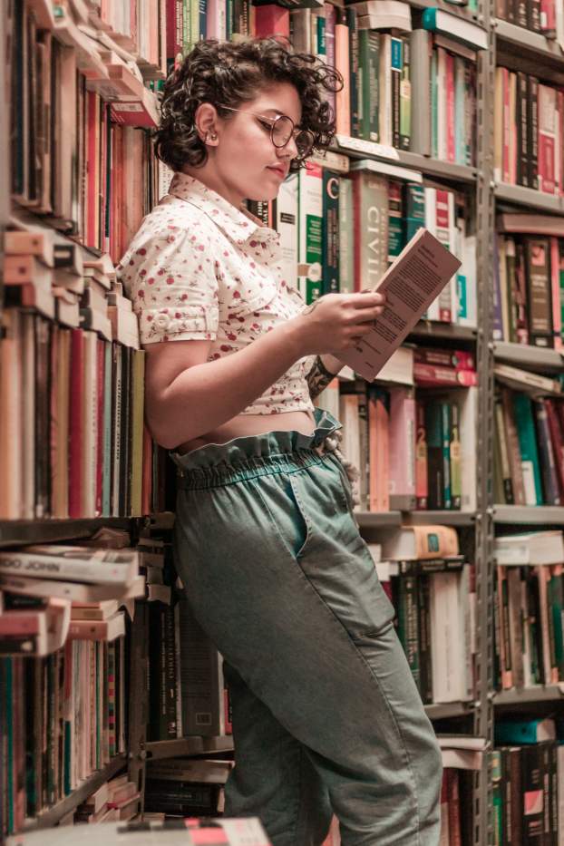 woman-holding-book-leaning-on-books-on-shelves-2167677.jpg