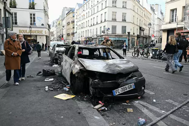 Egy a zavargások során felgyújtott autó kiégett roncsa mellett sétálnak emberek Párizsban, 2026 október 18-án.